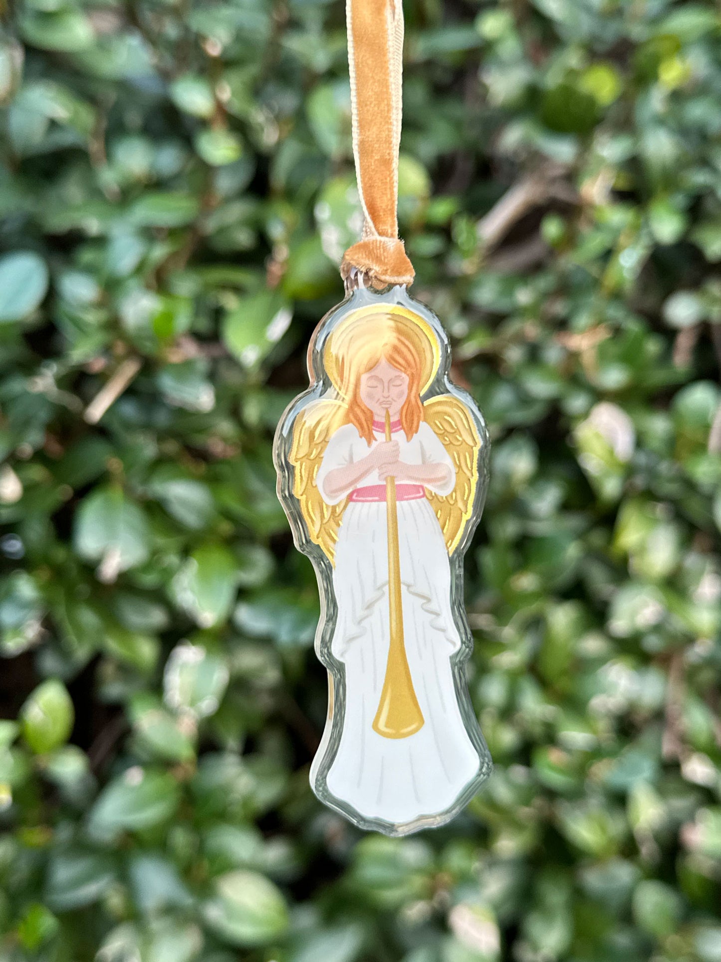 Angel ornament in white dress with pink sash blowing a horn with gold accents on the halo and wings hung from a velvet ribbon against a green plant background