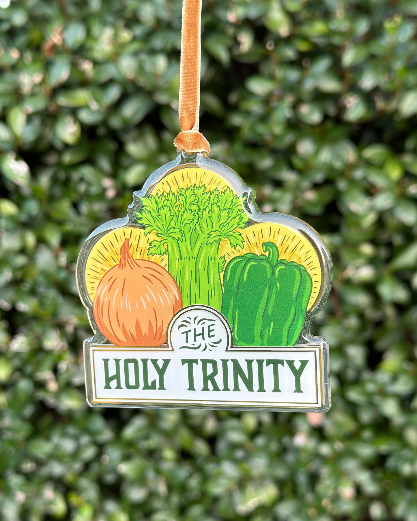 Decorative ornament with vegetables of onion, celery, and bell pepper and 'The Holy Trinity' text against a green leafy background hung from a velvet ribbon