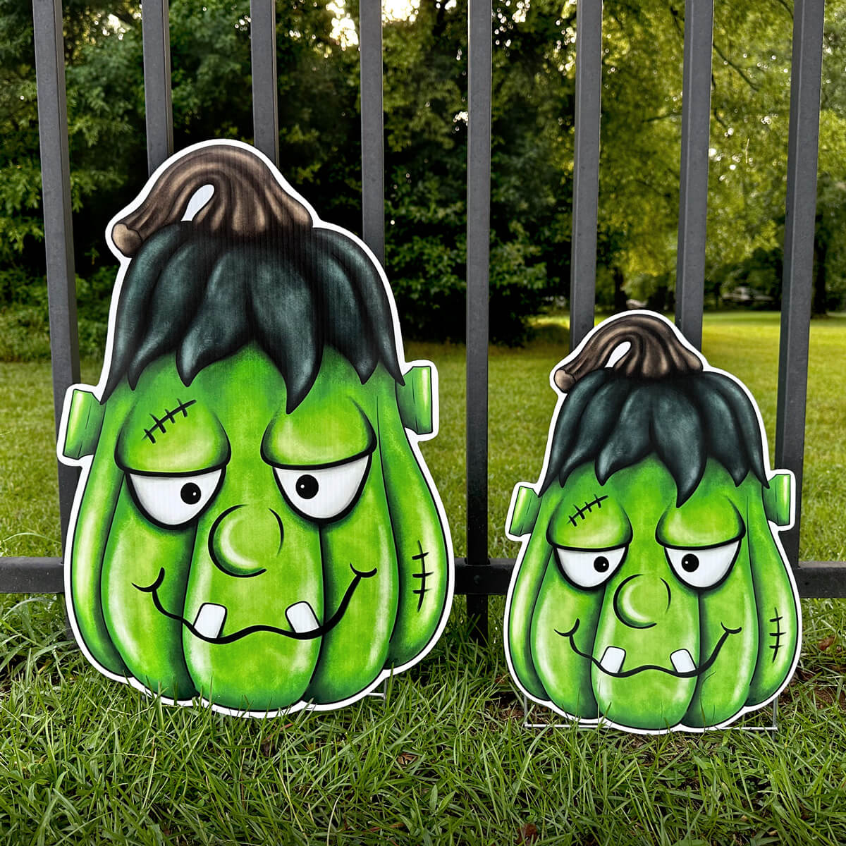 Two green Frankenstein-style pumpkin corrugated sign decorations on grass with a fence and trees in the background.
