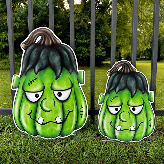 Two green Frankenstein-style pumpkin corrugated sign decorations on grass with a fence and trees in the background.