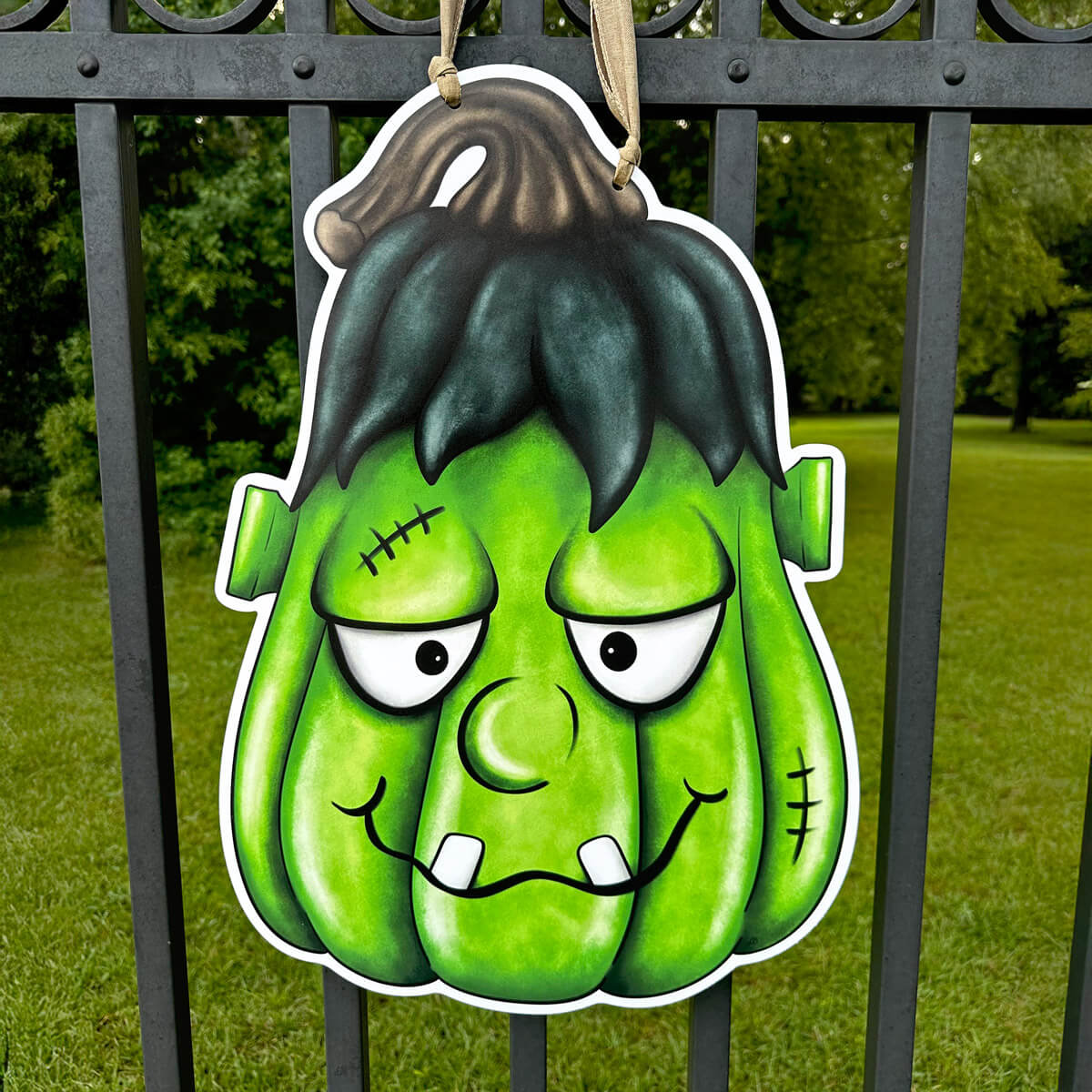 Halloween green monster Frankenstein inspired pumpkin head door hanger decoration hanging on a metal fence with grass and trees in the background.