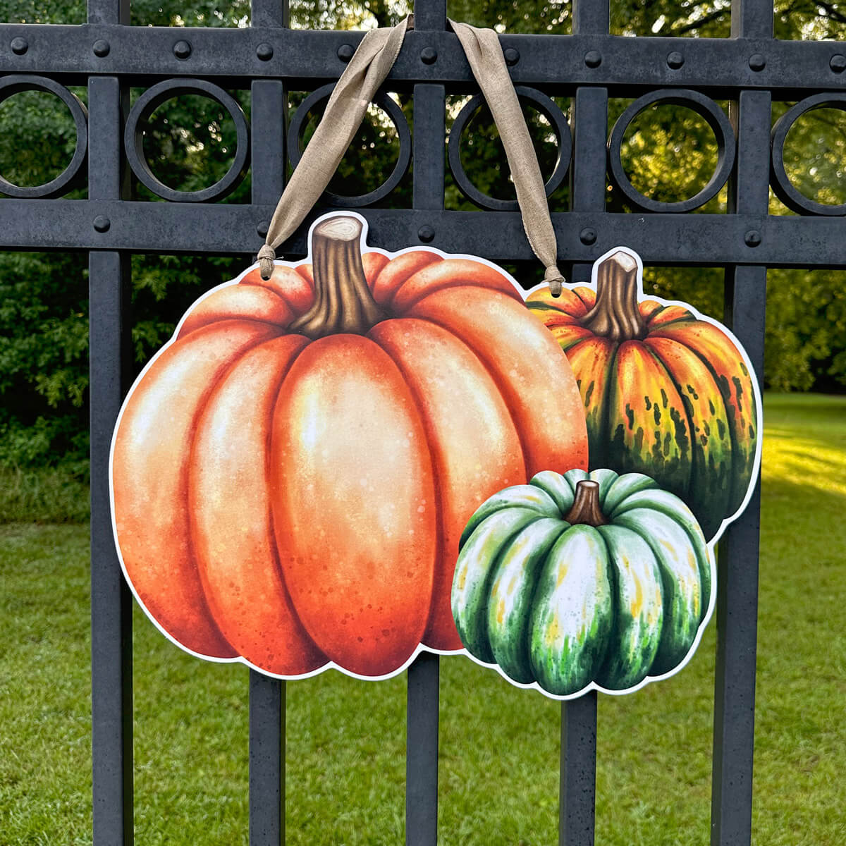 Decorative fall door hanger of pumpkins and gourds hanging on a metal fence with grass and trees in the background.
