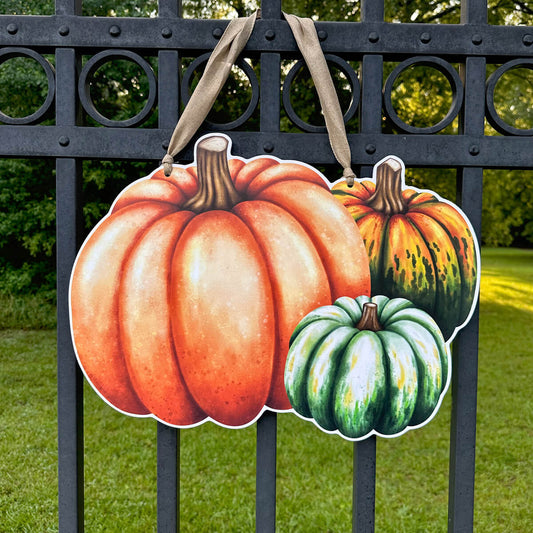 Decorative fall door hanger of pumpkins  and gourds hanging on a metal fence with grass and trees in the background.