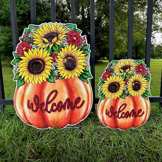 Two decorative, corrugated signs shaped like pumpkins with sunflowers and flowers, featuring the word 'Welcome', on a grassy background.