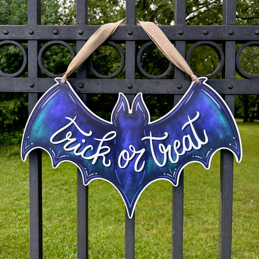 Deep purple bat-shaped door hanger with 'Trick or Treat' text hanging on a black metal gate.