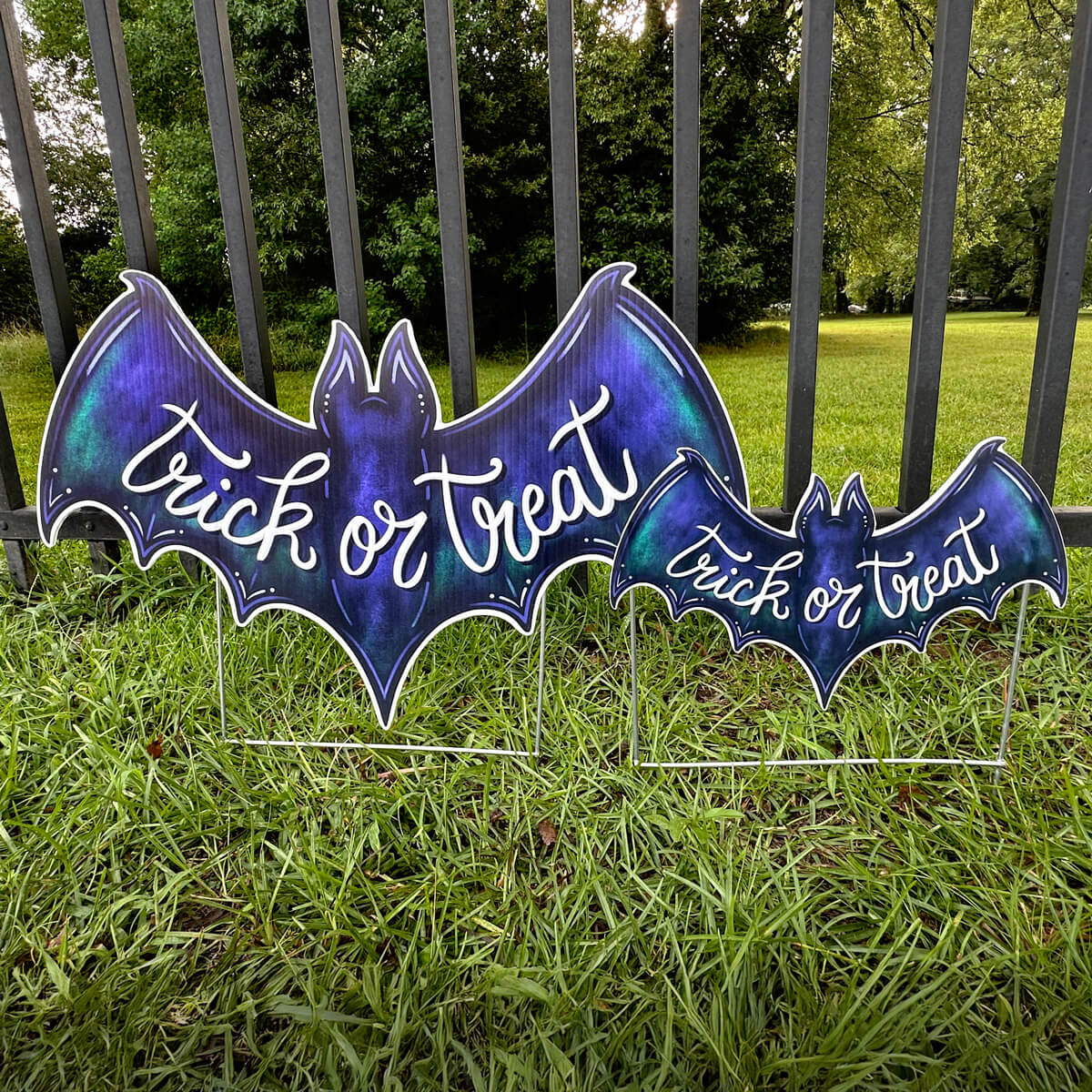 Two purple bat-shaped 'Trick or Treat' corrugated signs on grass with a fence and trees in the background.