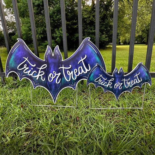 Two purple bat-shaped 'Trick or Treat' corrugated signs on grass with a fence and trees in the background.