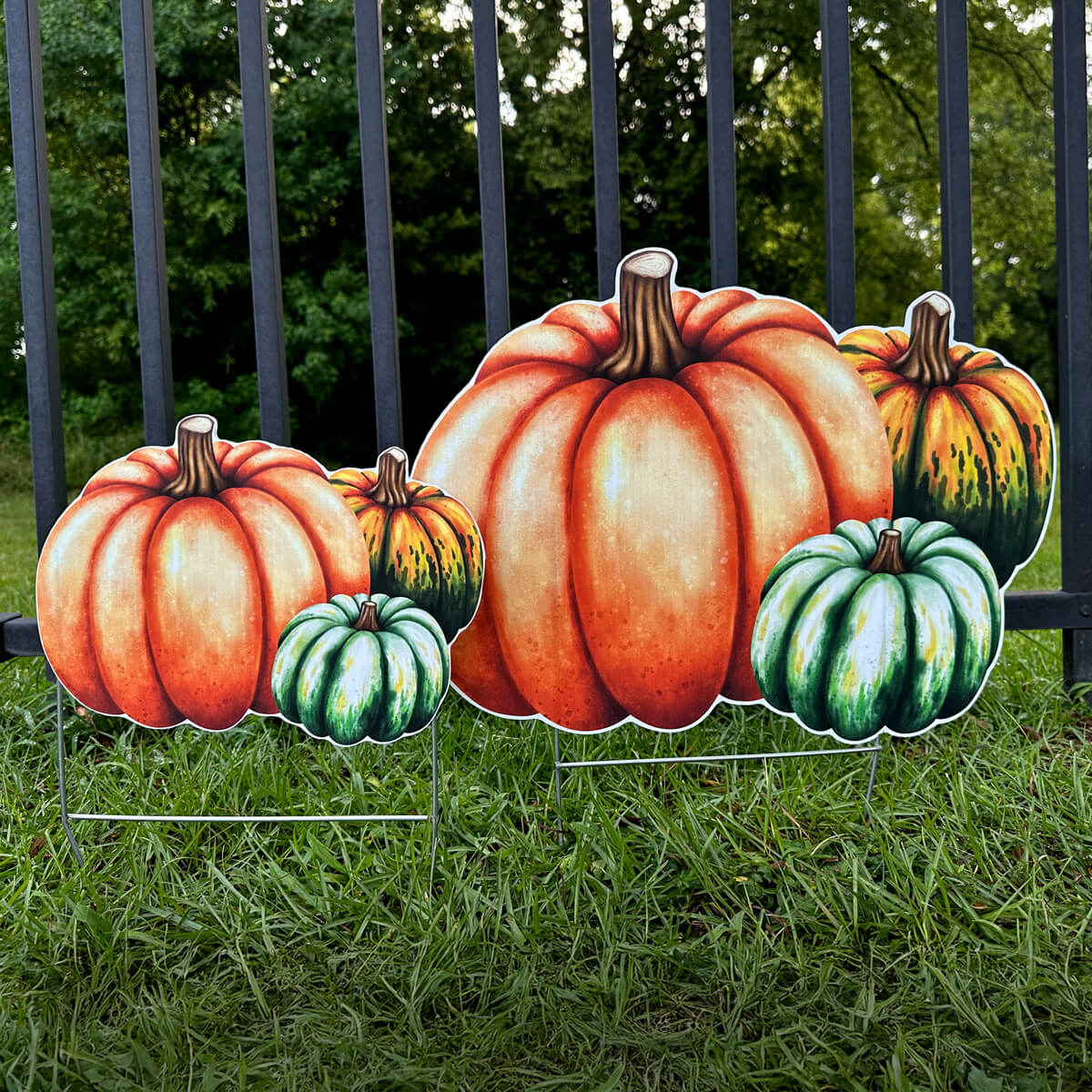 Fall decorative, corrugated pumpkin and gourd yard signs on grass with a fence and trees in the background.