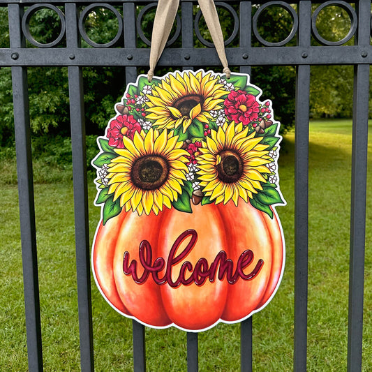 Decorative door hanger sign with sunflowers and flowers on a pumpkin, featuring the word 'Welcome', hanging on a metal fence.