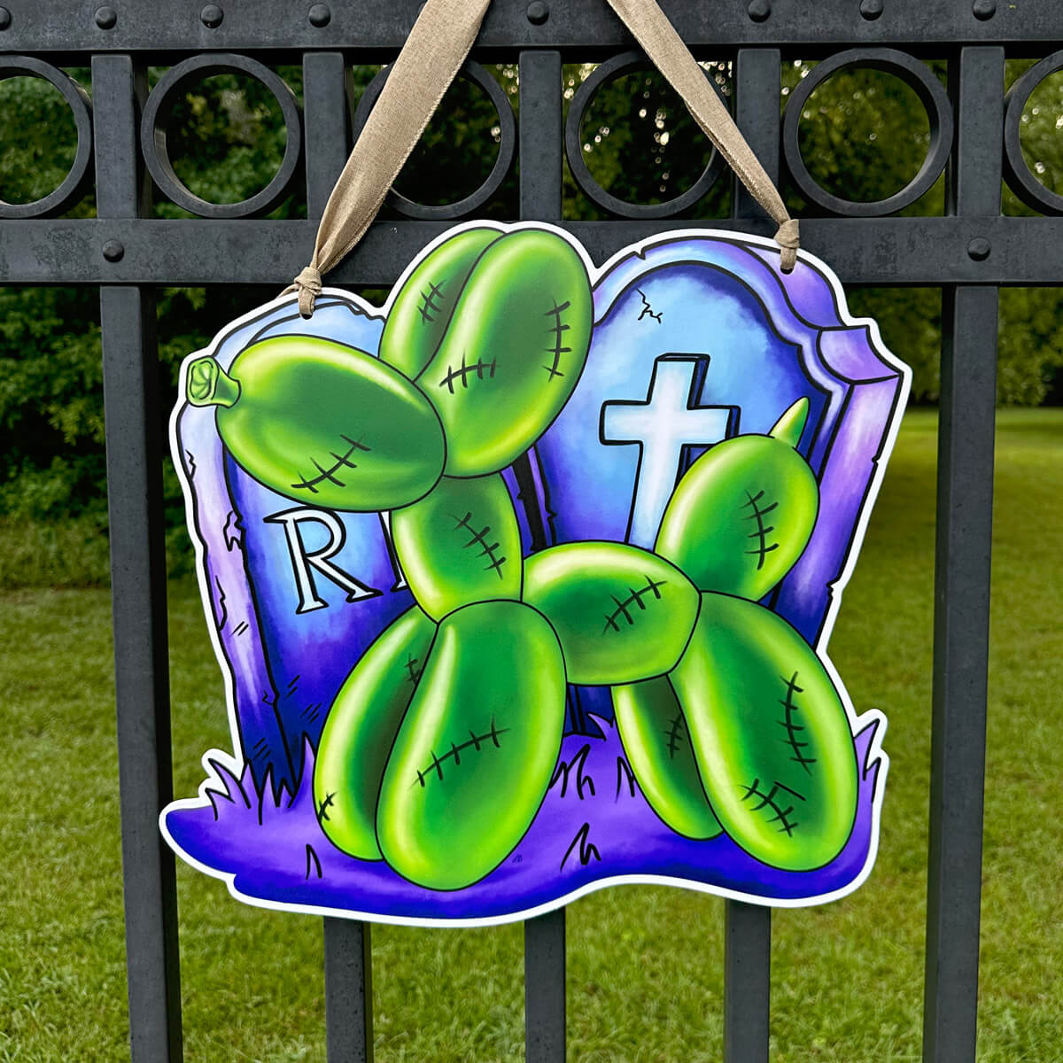 Decorative Halloween door hanger sign with green zombie balloon dog design in front of tombstones on a purple background, hanging on a metal fence.