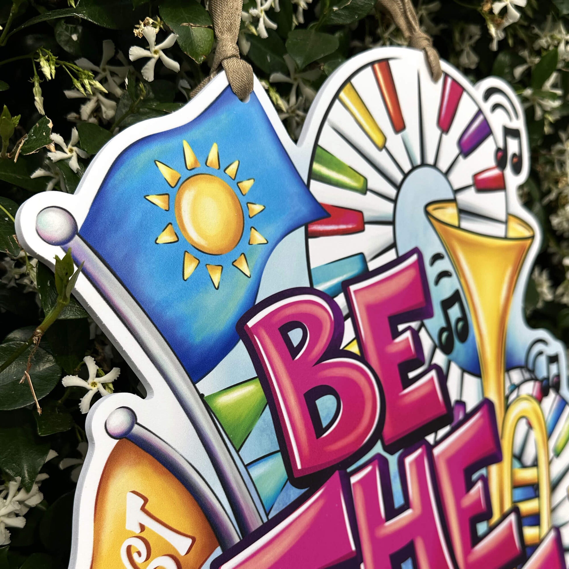 Decorative sign with colorful design and music notes and 'BE THE VIBE' text, hanging against a natural background.