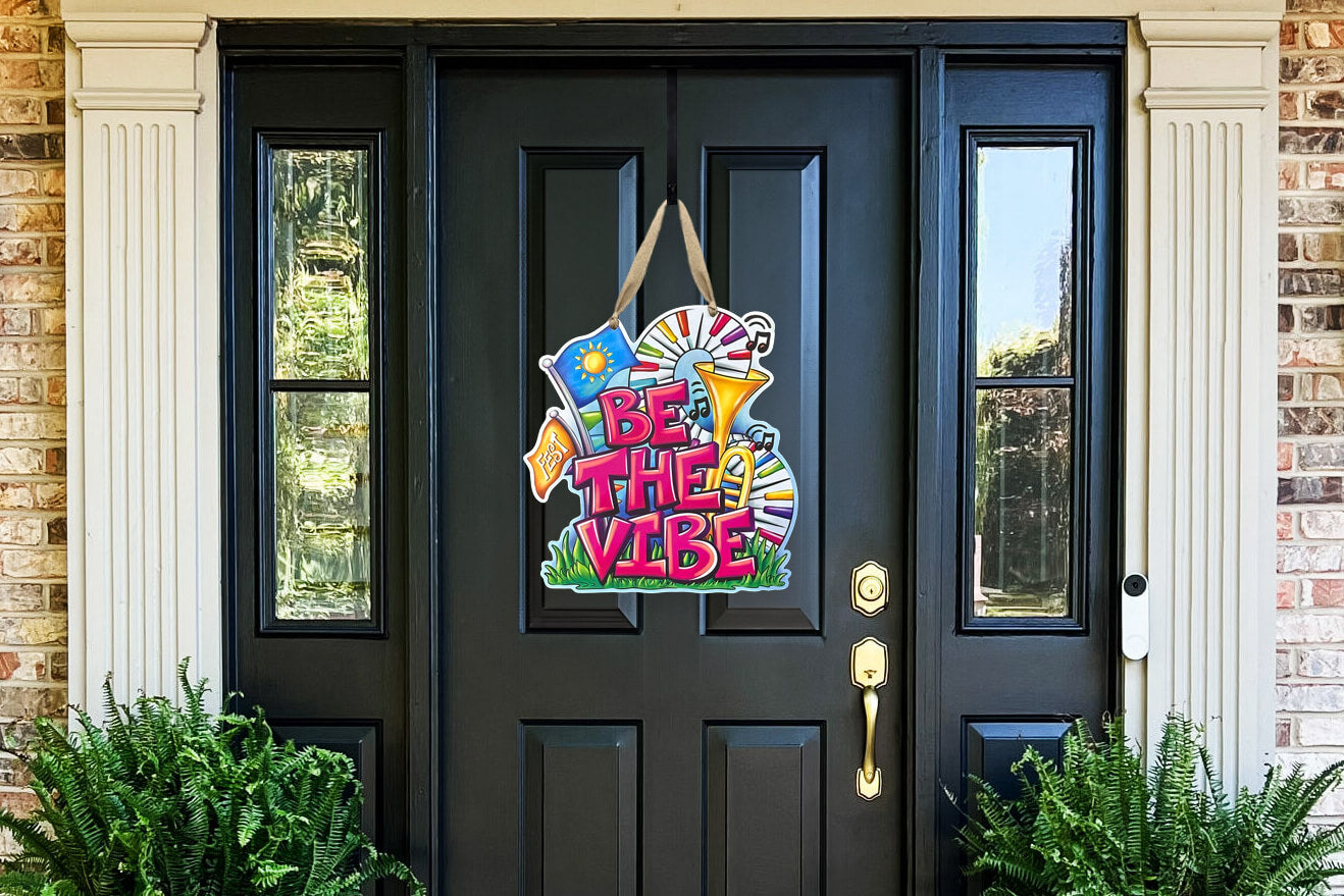Decorative and colorful door hanger on a black door with text 'Be the Vibe' in front of a brick building.