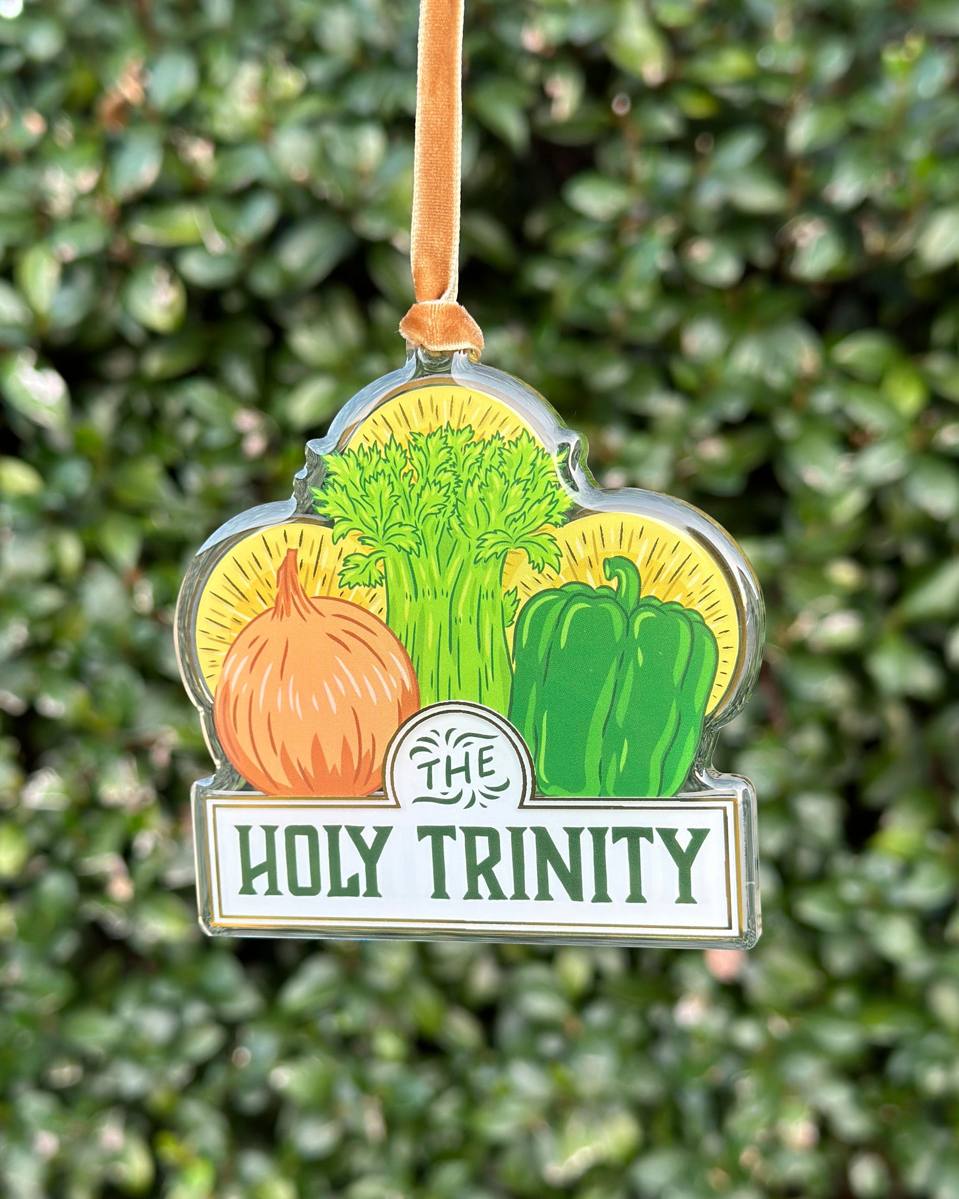 Decorative ornament with vegetables of onion, celery, and bell pepper and 'The Holy Trinity' text against a green leafy background hung from a velvet ribbon