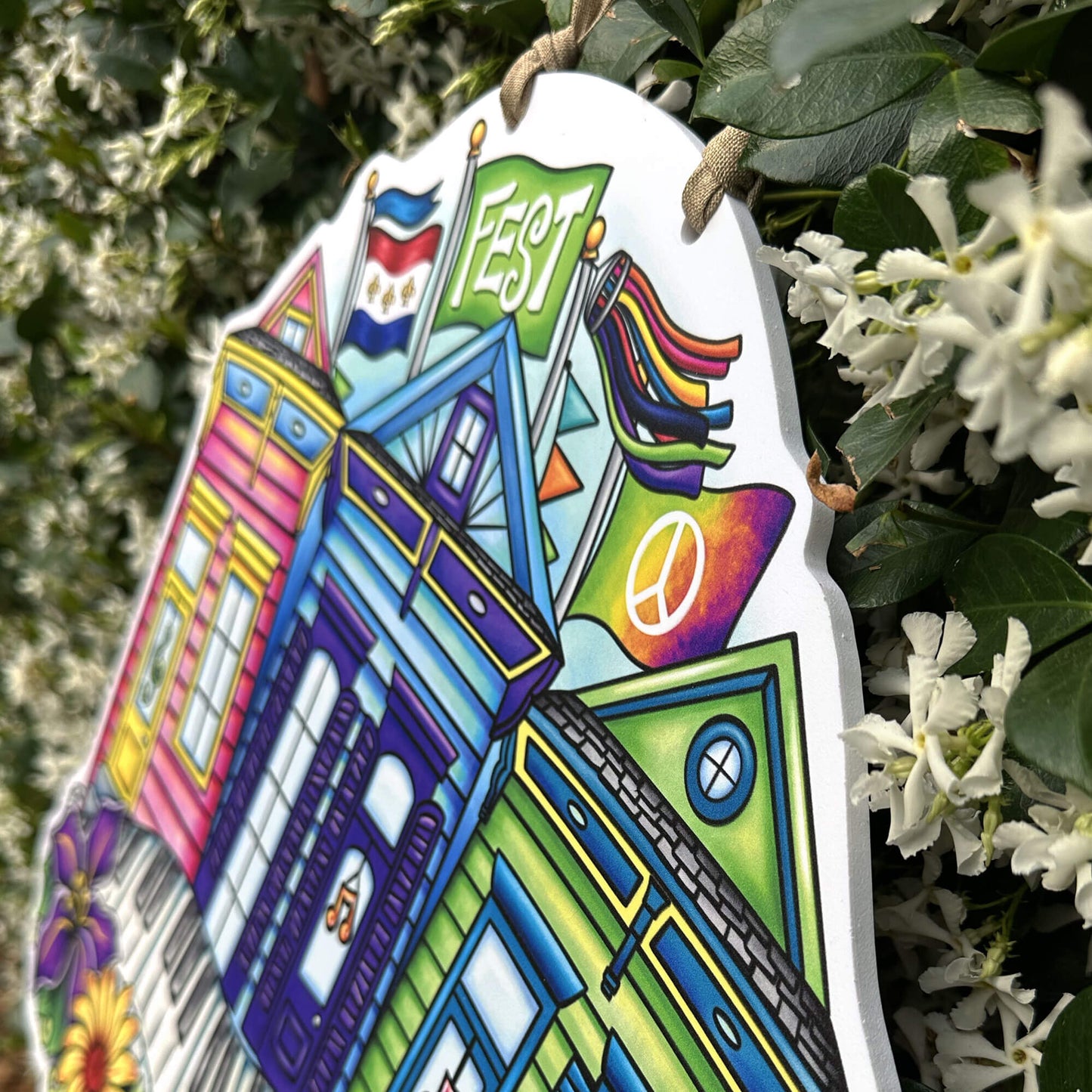 Colorful shotgun house-shaped sign with festival flags and symbols, hanging among greenery and flowers.