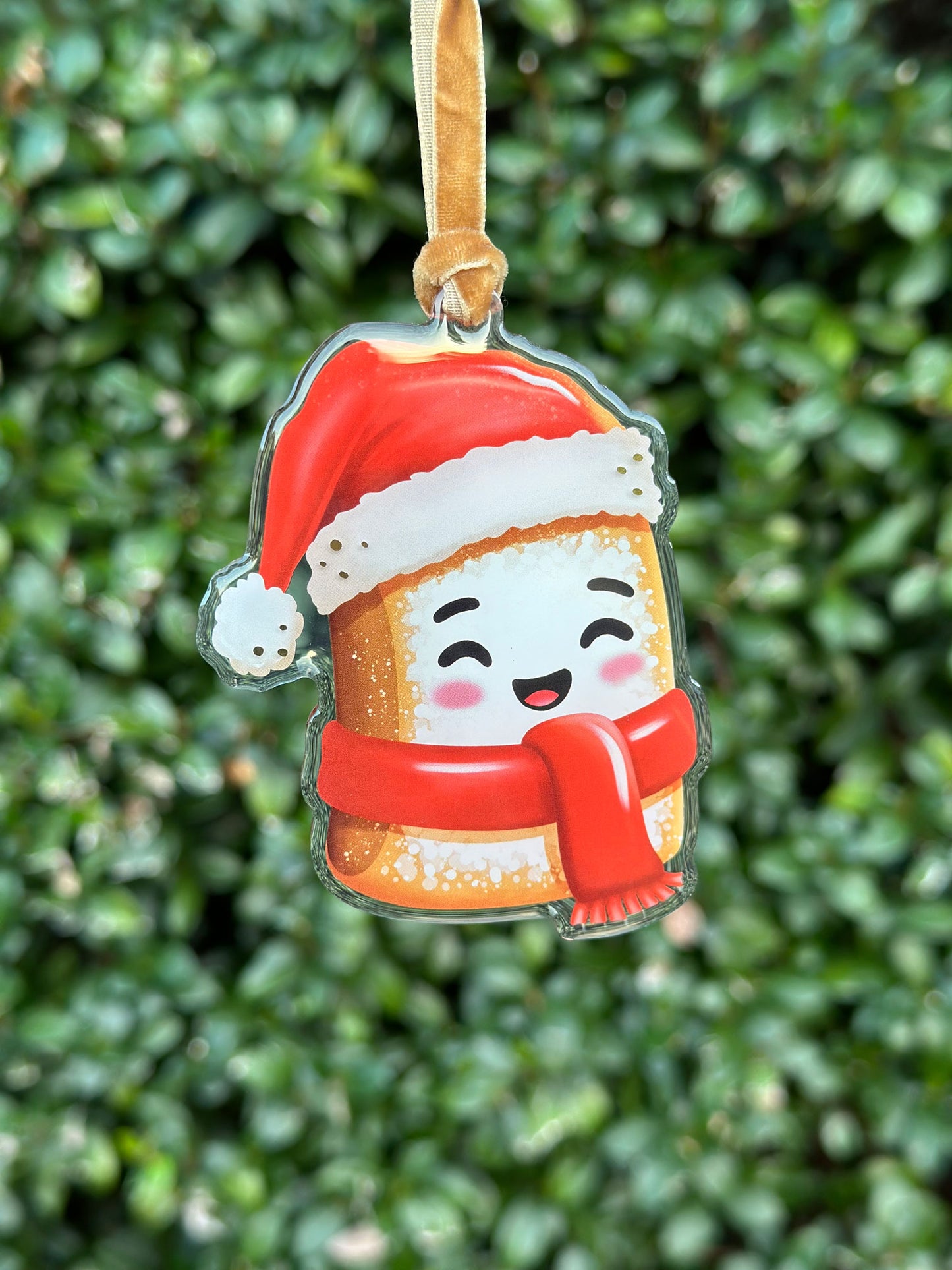 Decorative ornament shaped like a hot New Orleans beignet with a Santa hat and scarf, hanging from a velvet ribbon against a green leafy background.