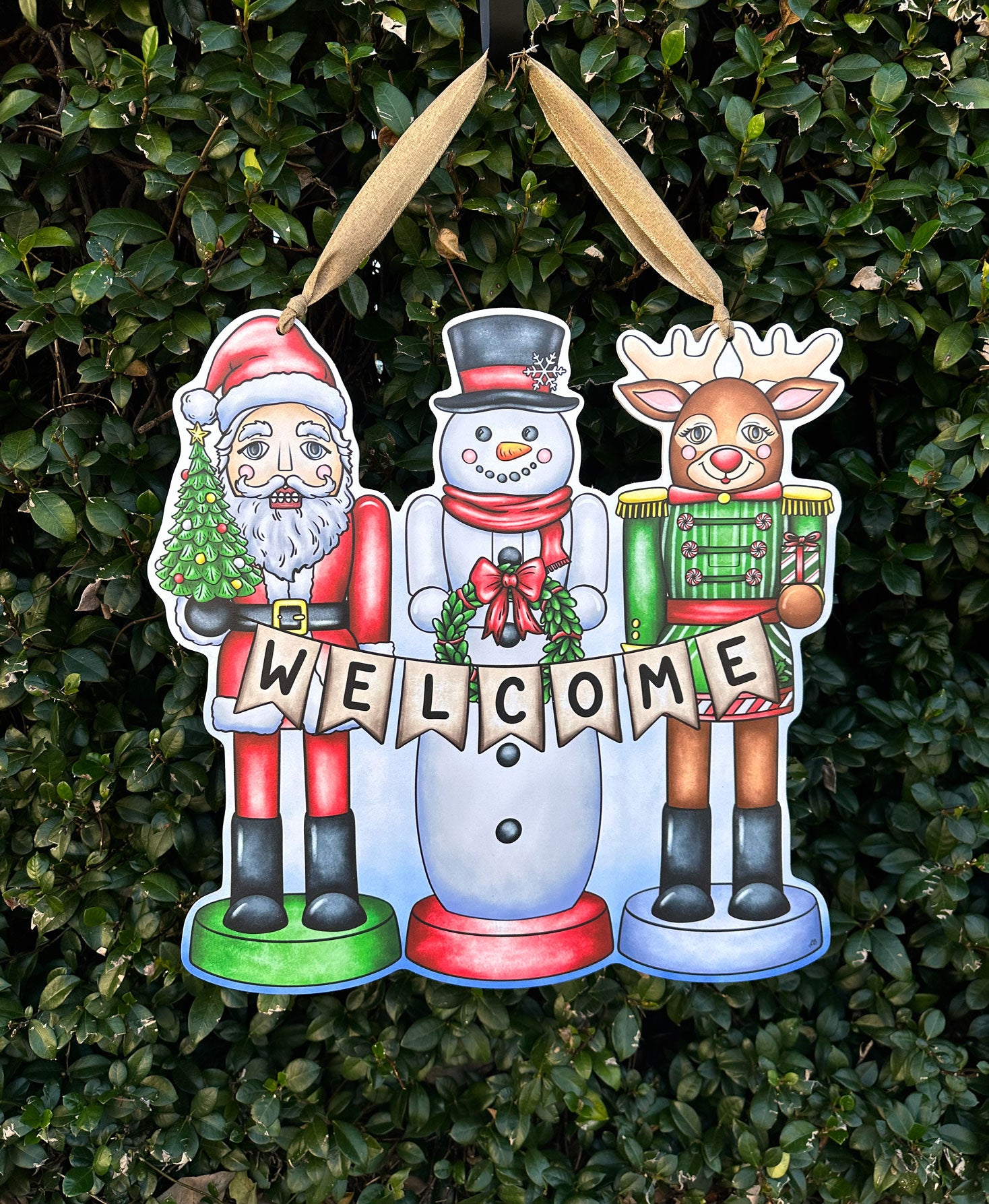 Decorative Christmas door decor with Santa, snowman, and reindeer holding a 'Welcome' sign against a green bush background.