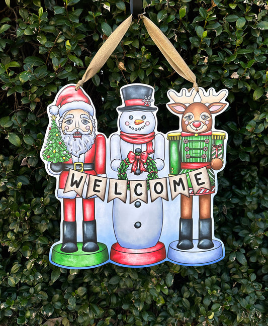 Decorative Christmas door decor with Santa, snowman, and reindeer holding a 'Welcome' sign against a green bush background.