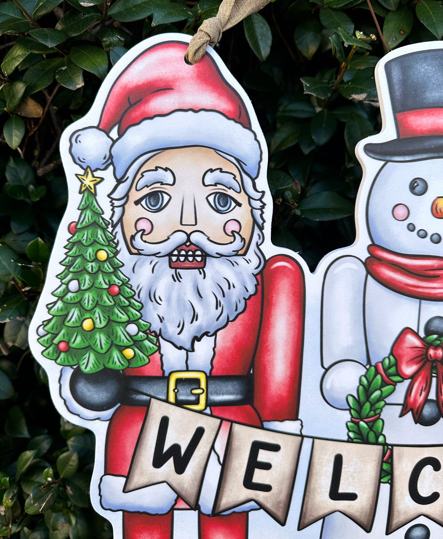 Decorative Christmas door decor detail of Santa Claus holding a tree and snowman against a green leafy background