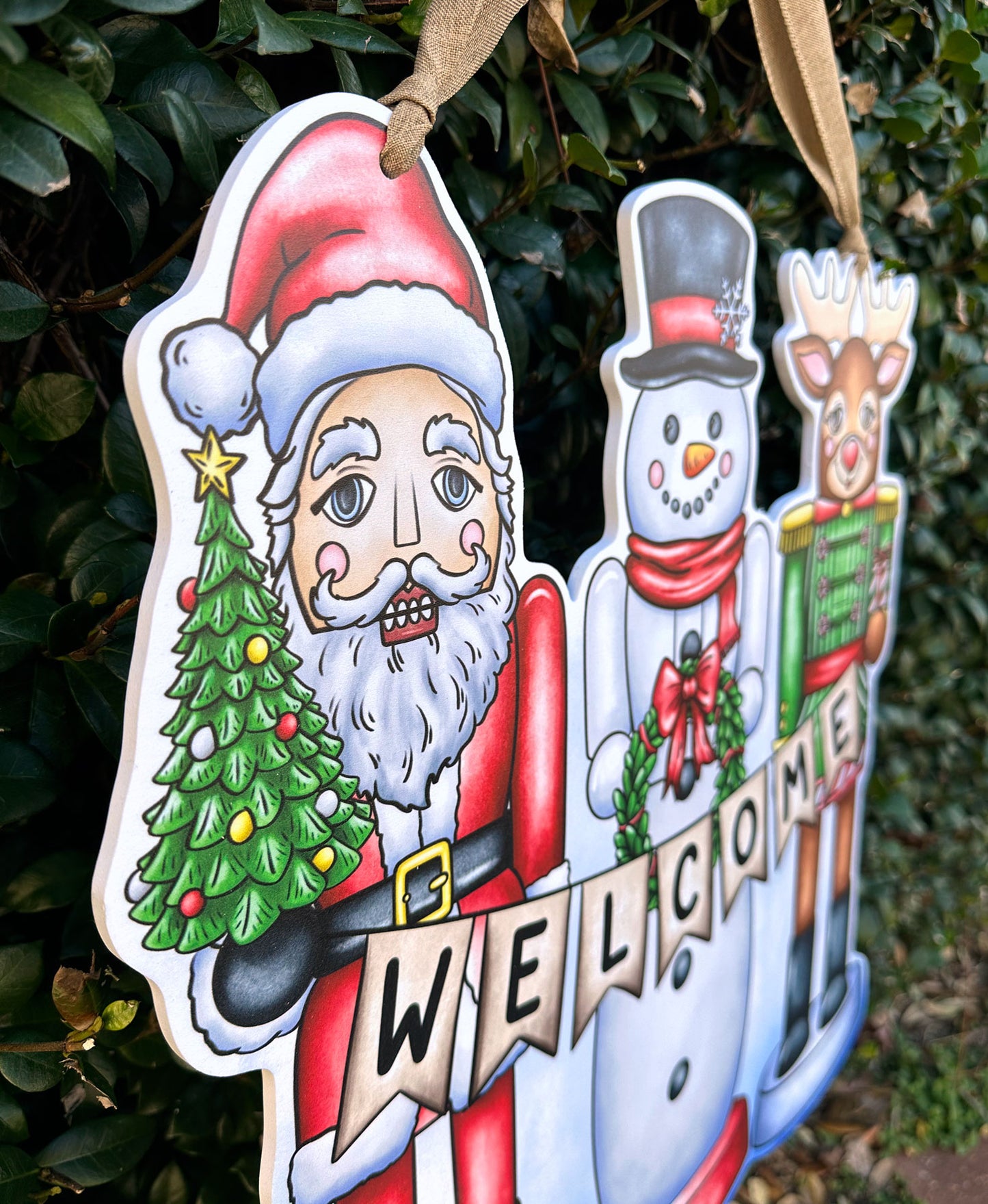 Side view of a decorative door hanger with Santa Claus, snowman, and reindeer against a green leafy background.
