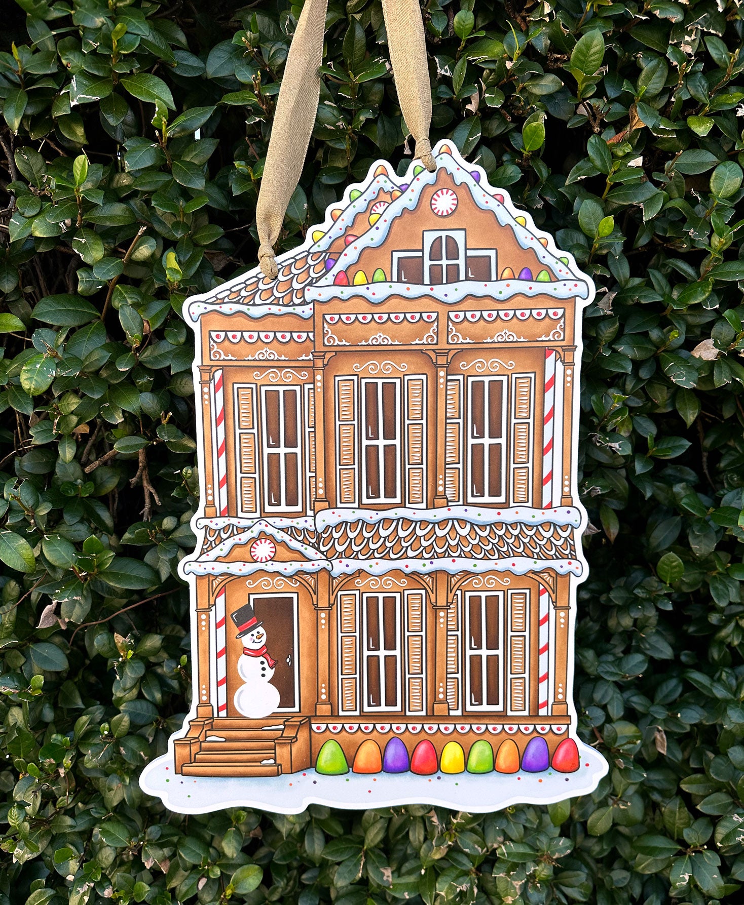 Decorative gingerbread shotgun house door decor hanging on a green leafy background