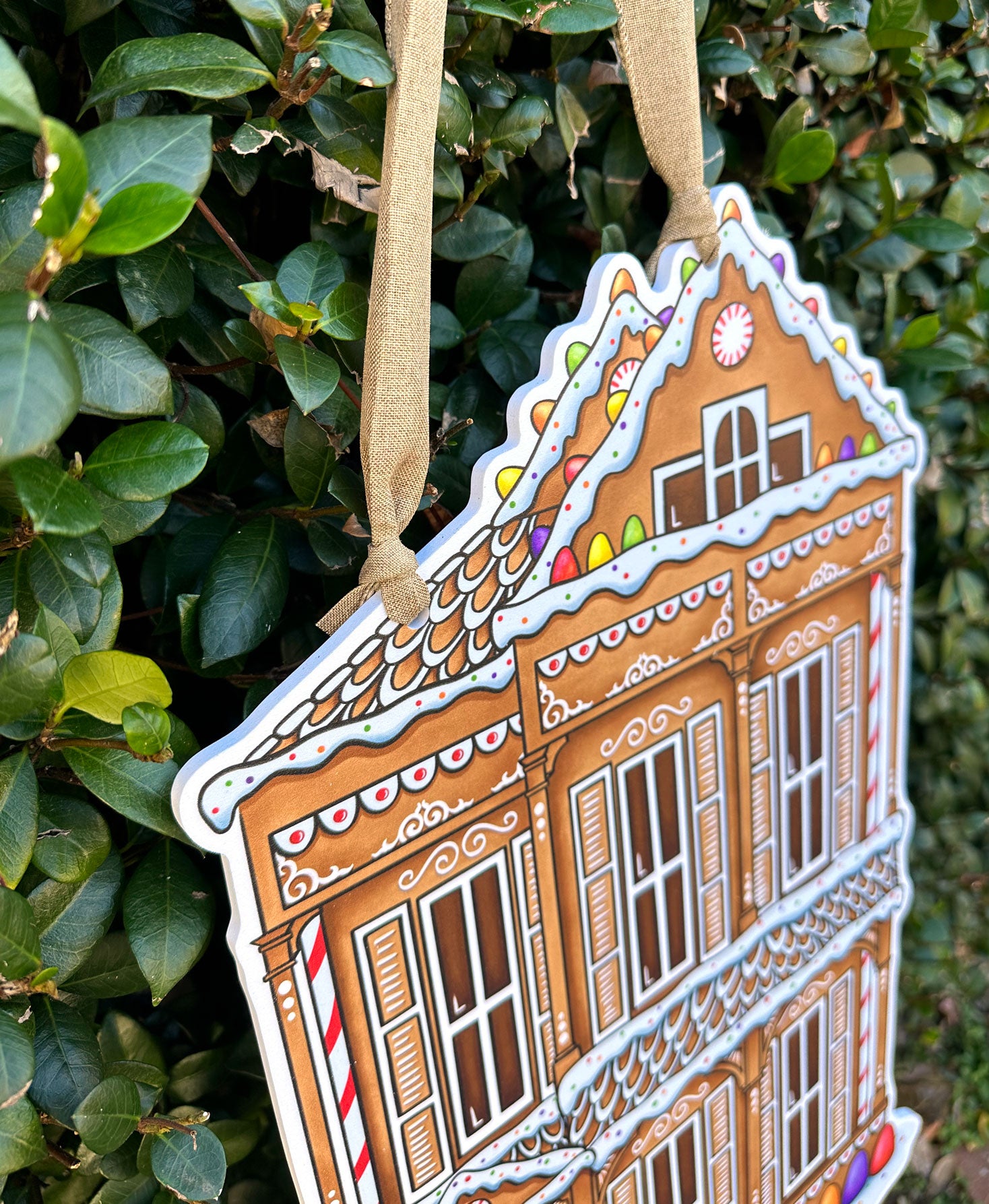 Details of a decorative gingerbread shotgun house door decor hanging against a green leafy background.