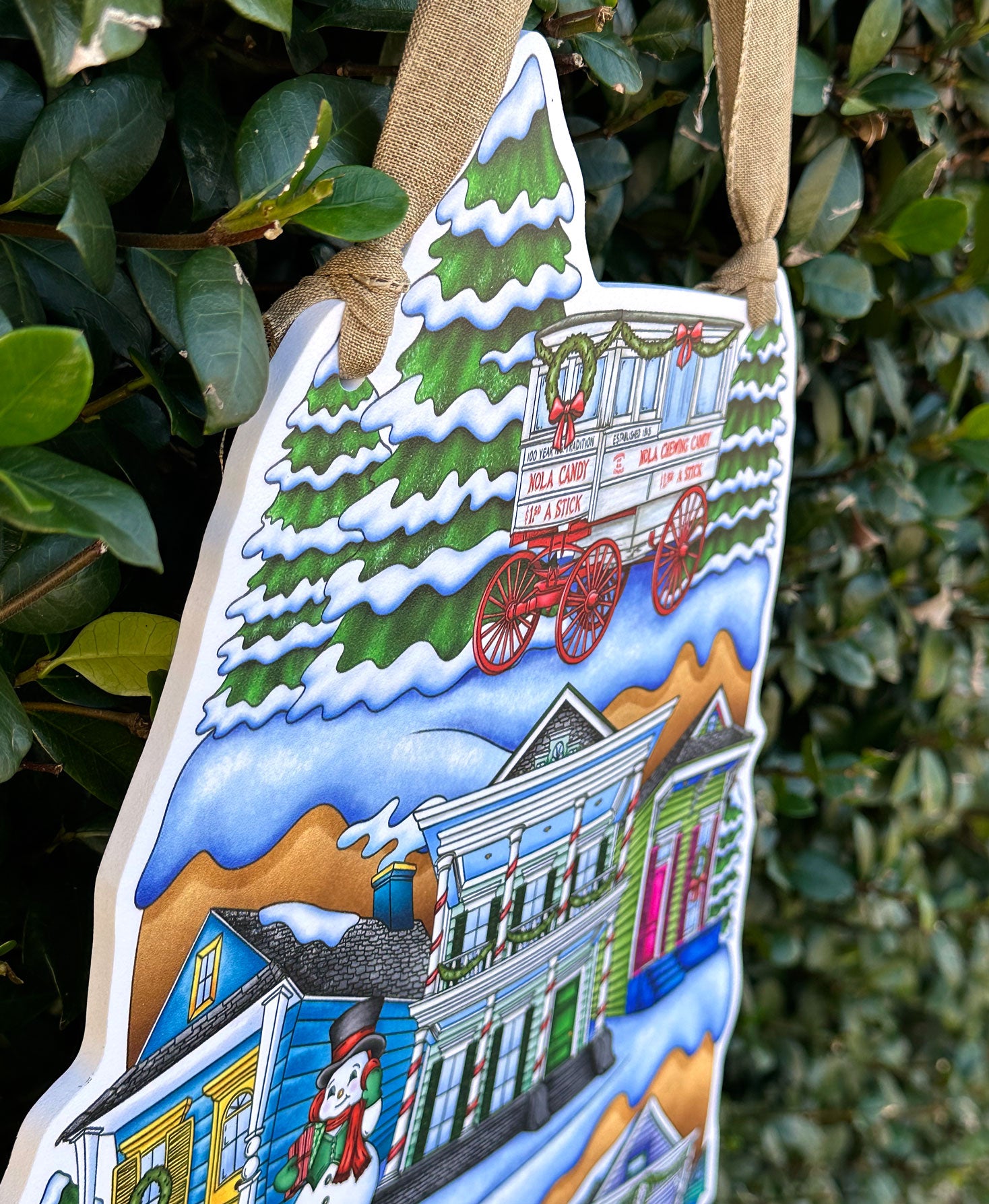 Detail of decorative Christmas door decor with a snowy village scene of colorful New Orleans shotgun homes on a green leafy background.