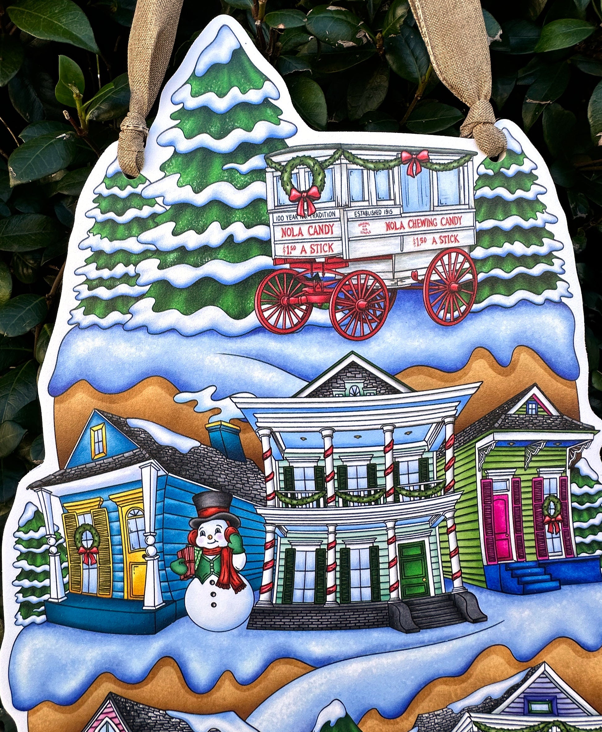 Decorative Christmas door decor close up with a snowy village scene of colorful New Orleans shotgun homes, a snowman, and candy cart on a green leafy background.