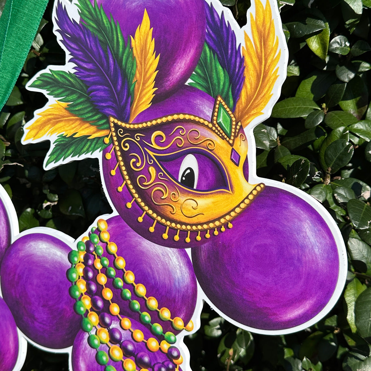 Close up of decorative Mardi Gras door hanger of a bead dog with purple, green, and gold tones. Bead dog is wearing beads and a mask with features against a green leafy background.