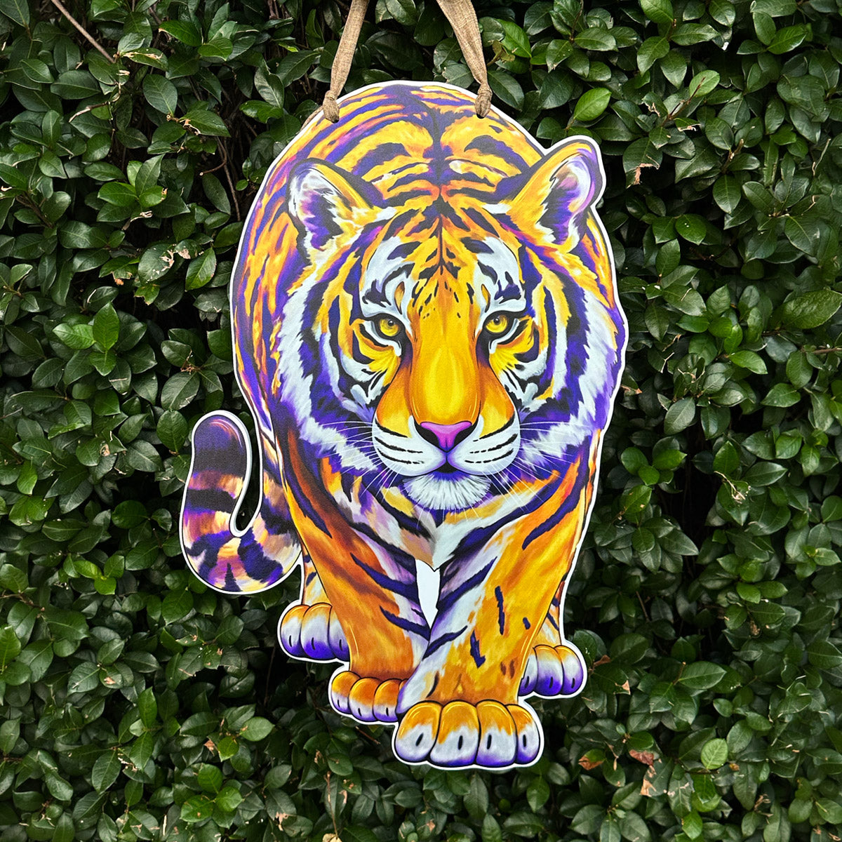 A hanging tiger-shaped decorative front door decor, featuring a colorful illustration of a tiger in orange, yellow, purple, and white, suspended by a tan ribbon against a background of dense green foliage.