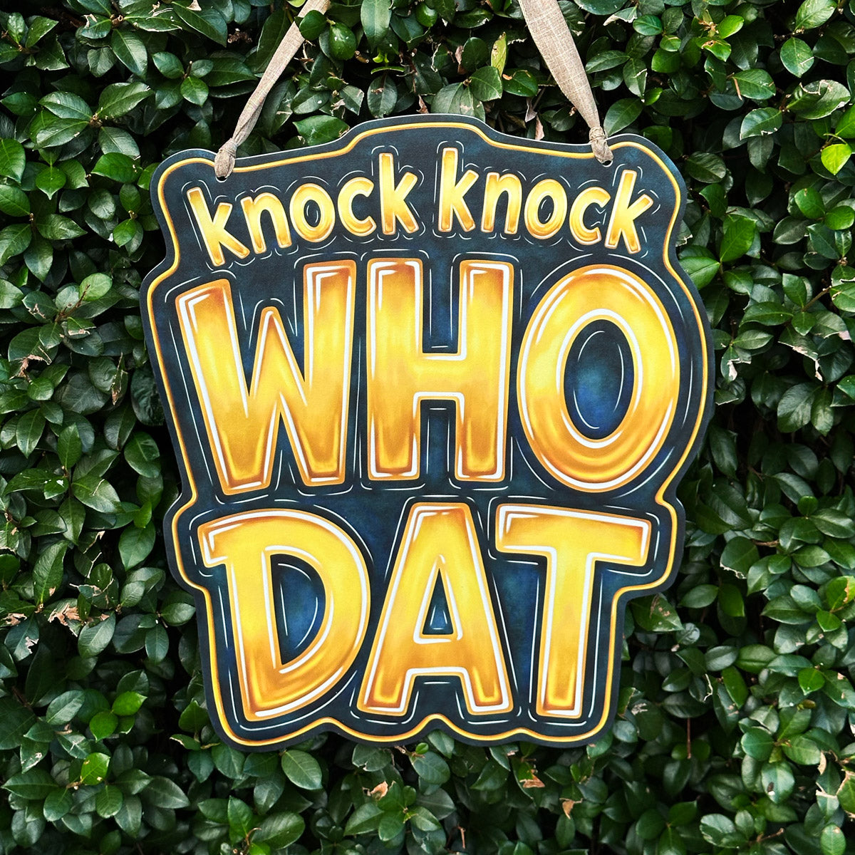 A charming door hanger with the phrase 'knock knock WHO DAT' in large gold letters outlined in blue and black tone, suspended against a background of dense green foliage.
